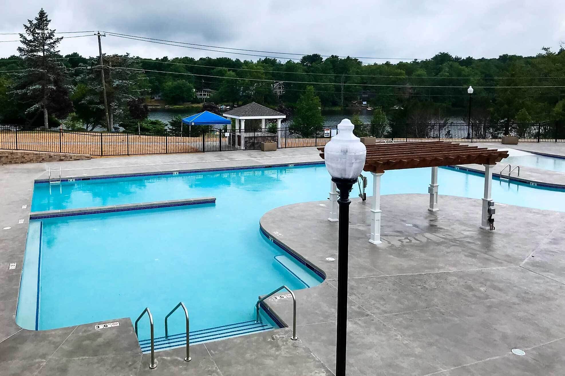 A swimming pool with clear blue water, surrounded by a concrete deck. There are metal railings along the pool's edge and a covered seating area with a pergola.