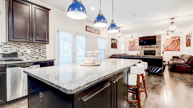 A modern kitchen with granite countertops, stainless steel appliances, and dark cabinetry. There are pendant lights, a dining area with a table and chairs, and a living room with a fireplace and a sofa. - Downtown DC Home w/ Furnished Patio! (Washington)
