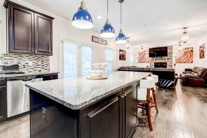 Une cuisine moderne dotée de plans de travail en granit, d’appareils électroménagers en acier inoxydable et de meubles de couleur sombre. On y trouve des luminaires suspendus, un coin repas avec une table et des chaises, ainsi qu’un salon avec une cheminée et un canapé.