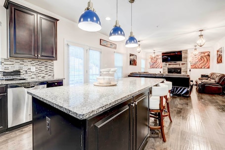 A modern kitchen with granite countertops, stainless steel appliances, and dark cabinetry. There are pendant lights, a dining area with a table and chairs, and a living room with a fireplace and a large TV.
