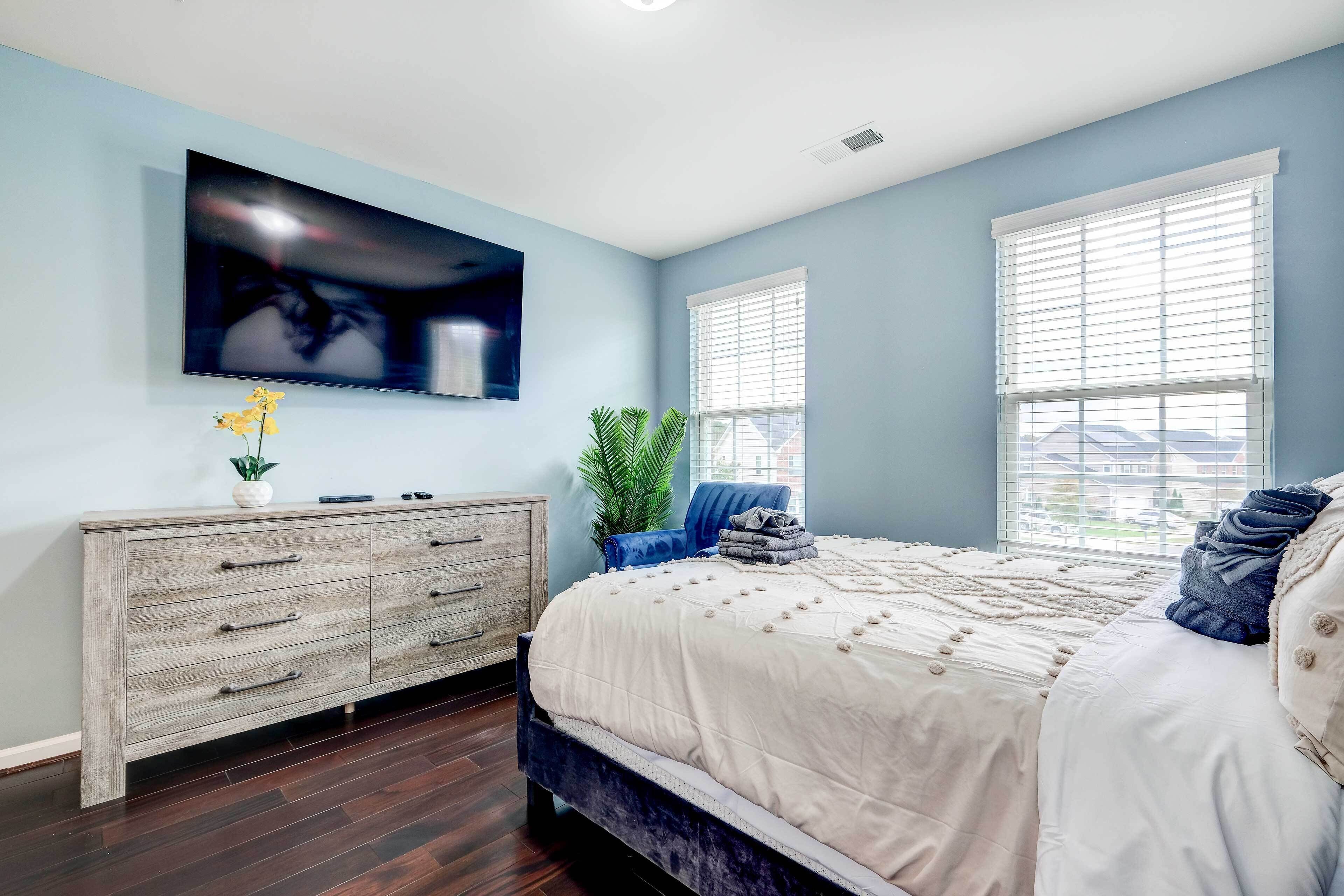 Une chambre à coucher comprenant un lit, un téléviseur fixé au mur, une commode et une fenêtre avec des stores.