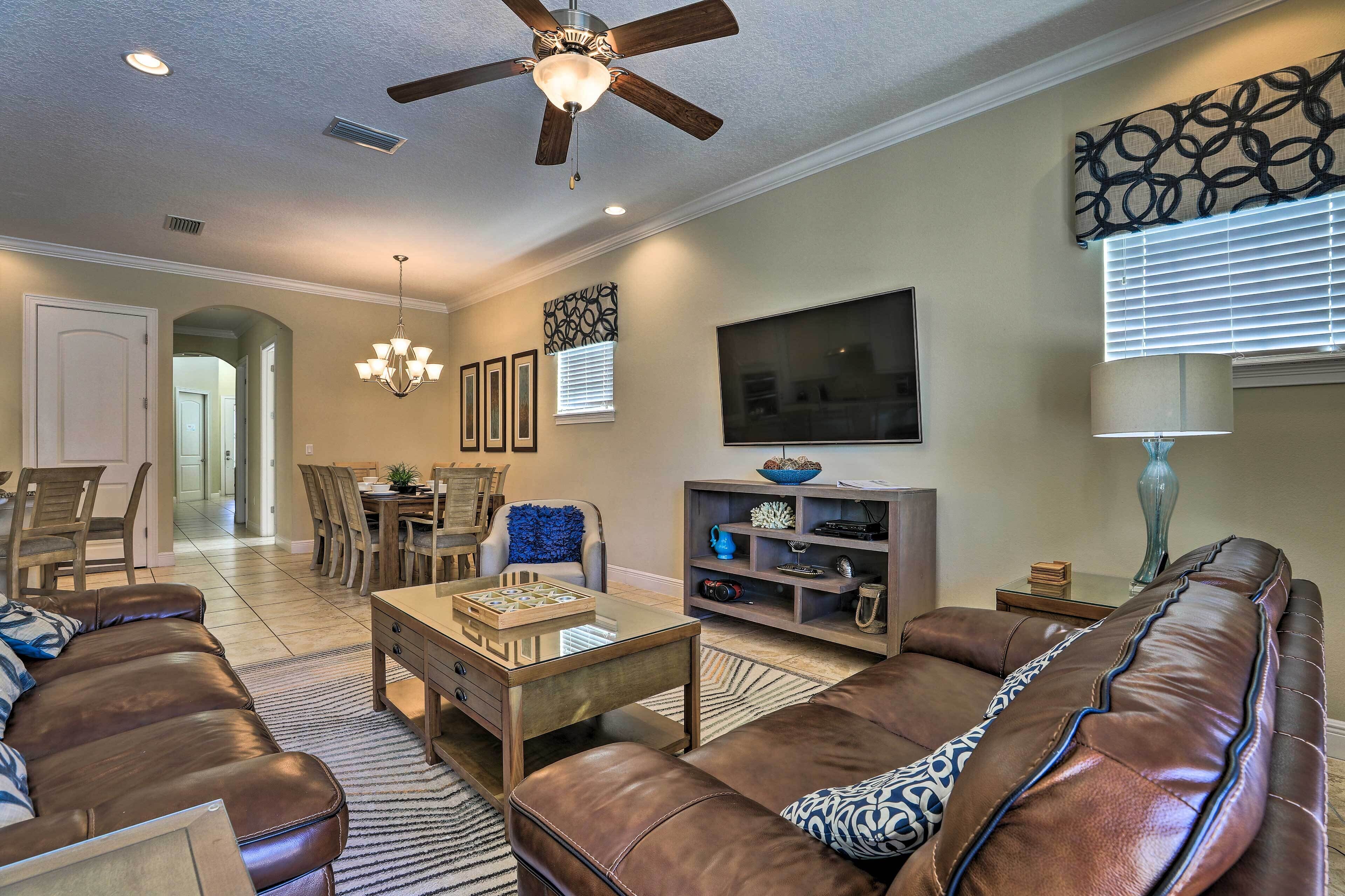 A living room with a brown leather sofa, a glass coffee table, a chandelier, a ceiling fan, and a television on the wall.