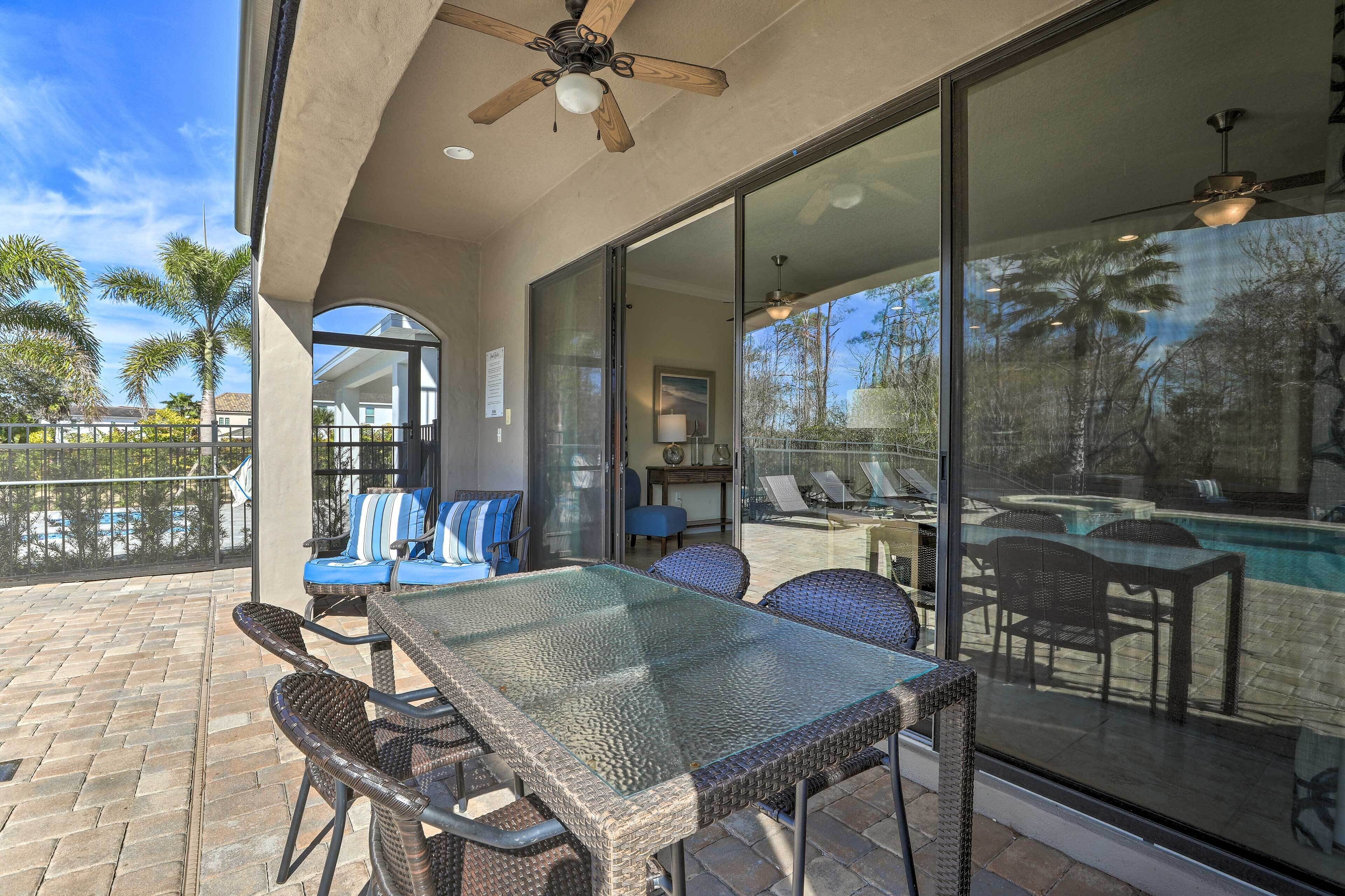 An outdoor patio with a glass table and chairs, a ceiling fan, and a view of a swimming pool.
