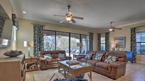 A spacious living room with leather sofas, a glass coffee table, and a ceiling fan. Large windows provide ample natural light and views of the outdoors. - Luxe Reunion House w/ Private Pool: Near Disney! (Kissimmee)