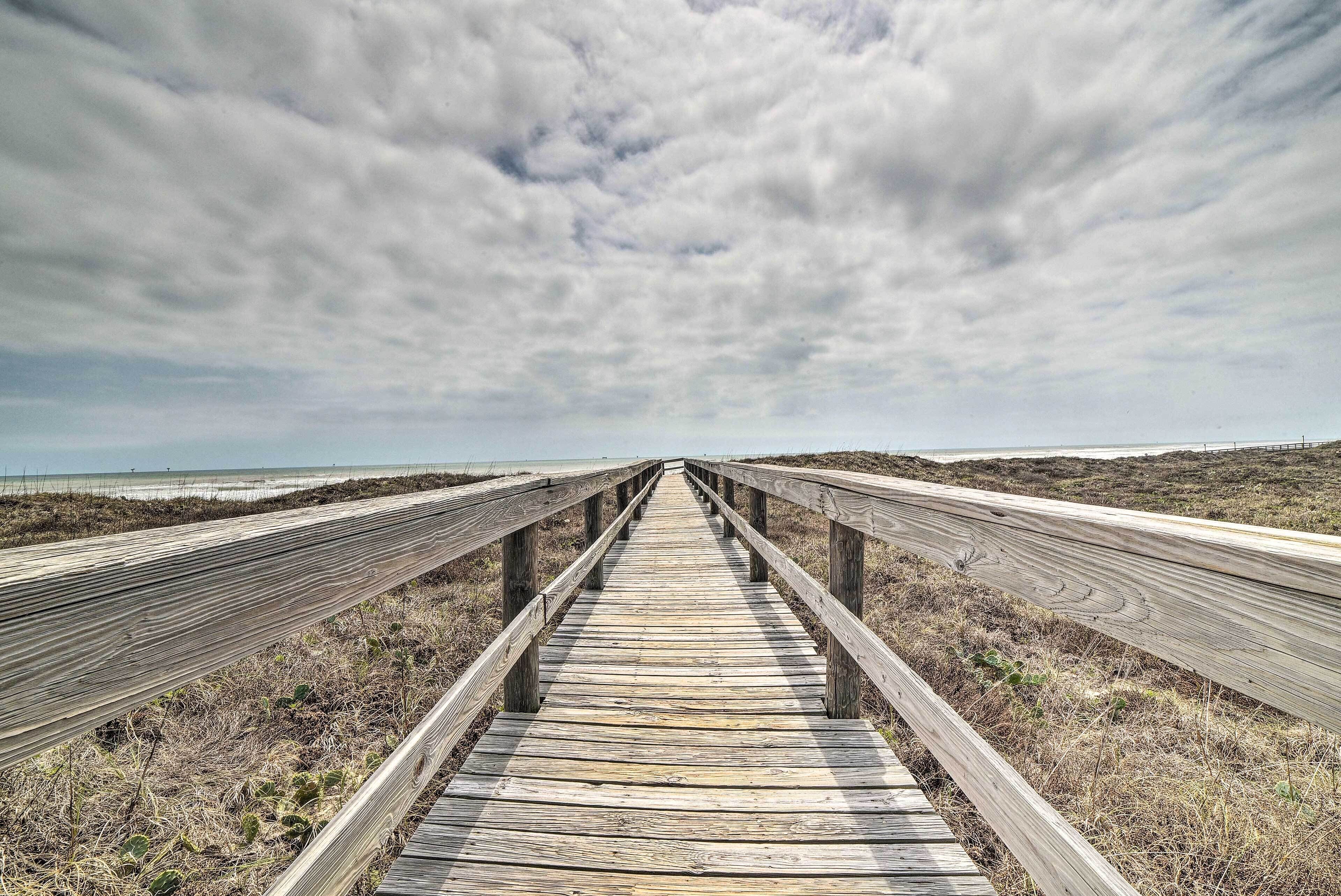 Un paseo marítimo de madera que conduce a la playa, rodeado de hierba seca y bajo un cielo nublado.
