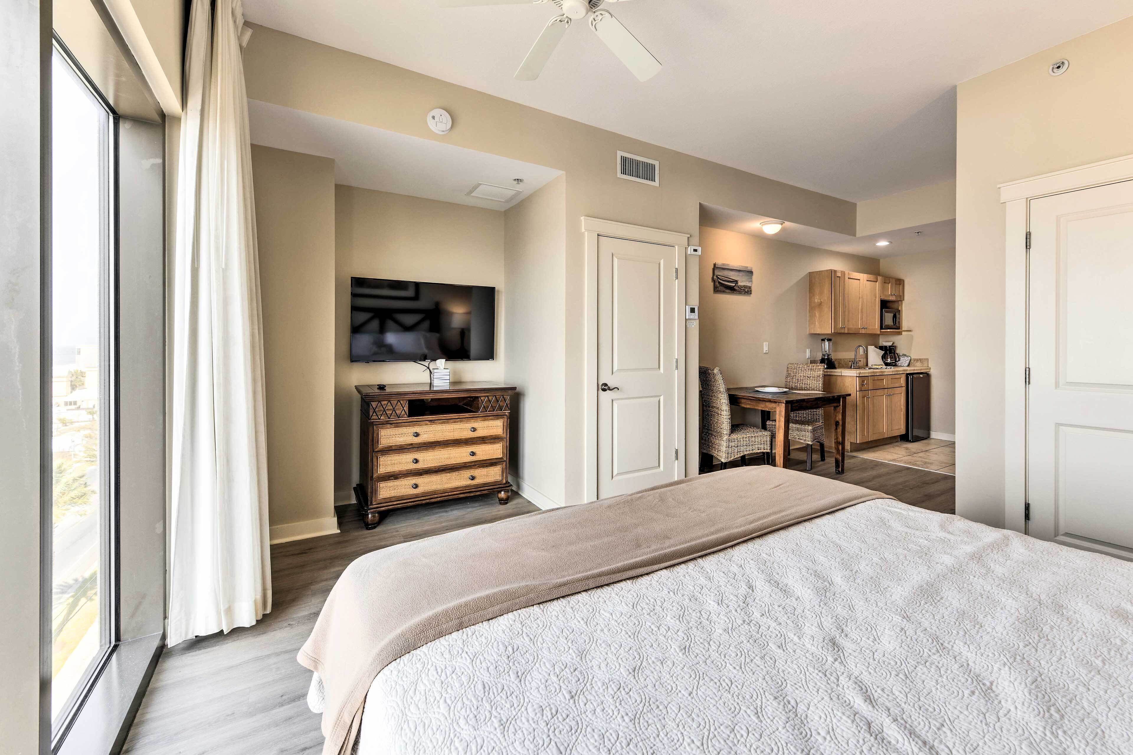 Un dormitorio con una cama grande, un televisor en la pared, una kitchenette y una puerta que da a otra habitación.