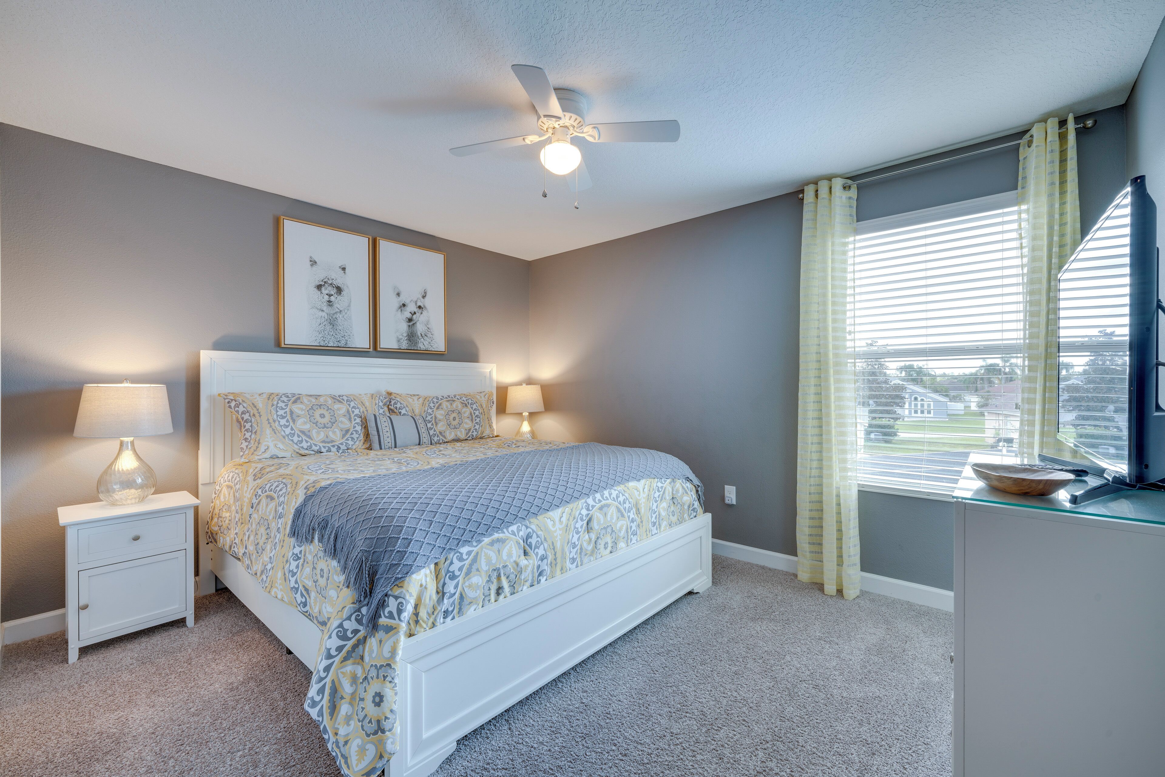 A bedroom with a bed, nightstand, lamp, ceiling fan, window with curtains, and a view of the outside.