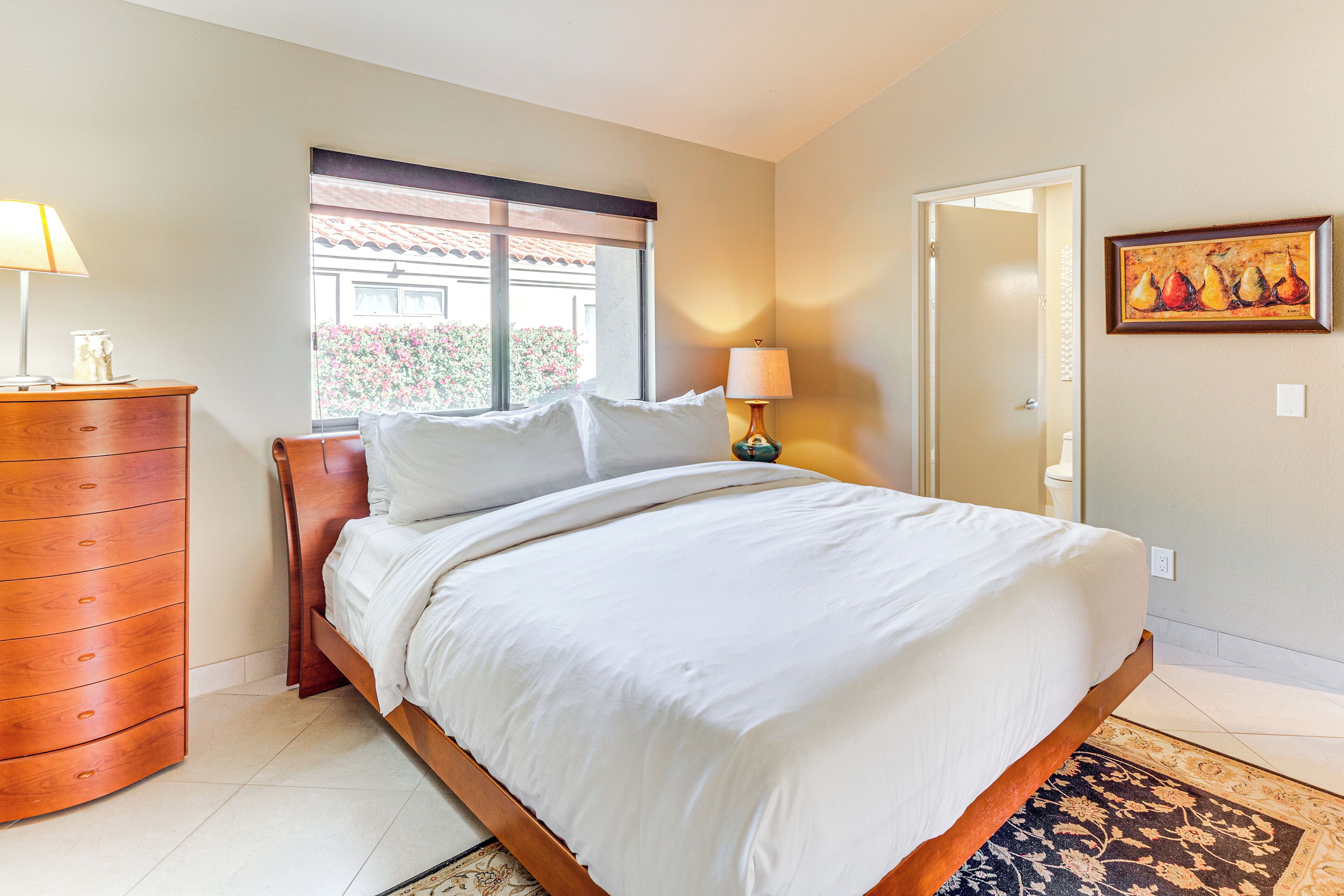 A bedroom with a bed, a wooden dresser, a window with a view, a door, and a painting on the wall.