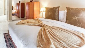 Un lit bien fait, recouvert d’une couverture beige, une armoire en bois et une lampe de chevet dans une chambre d’hôtel.