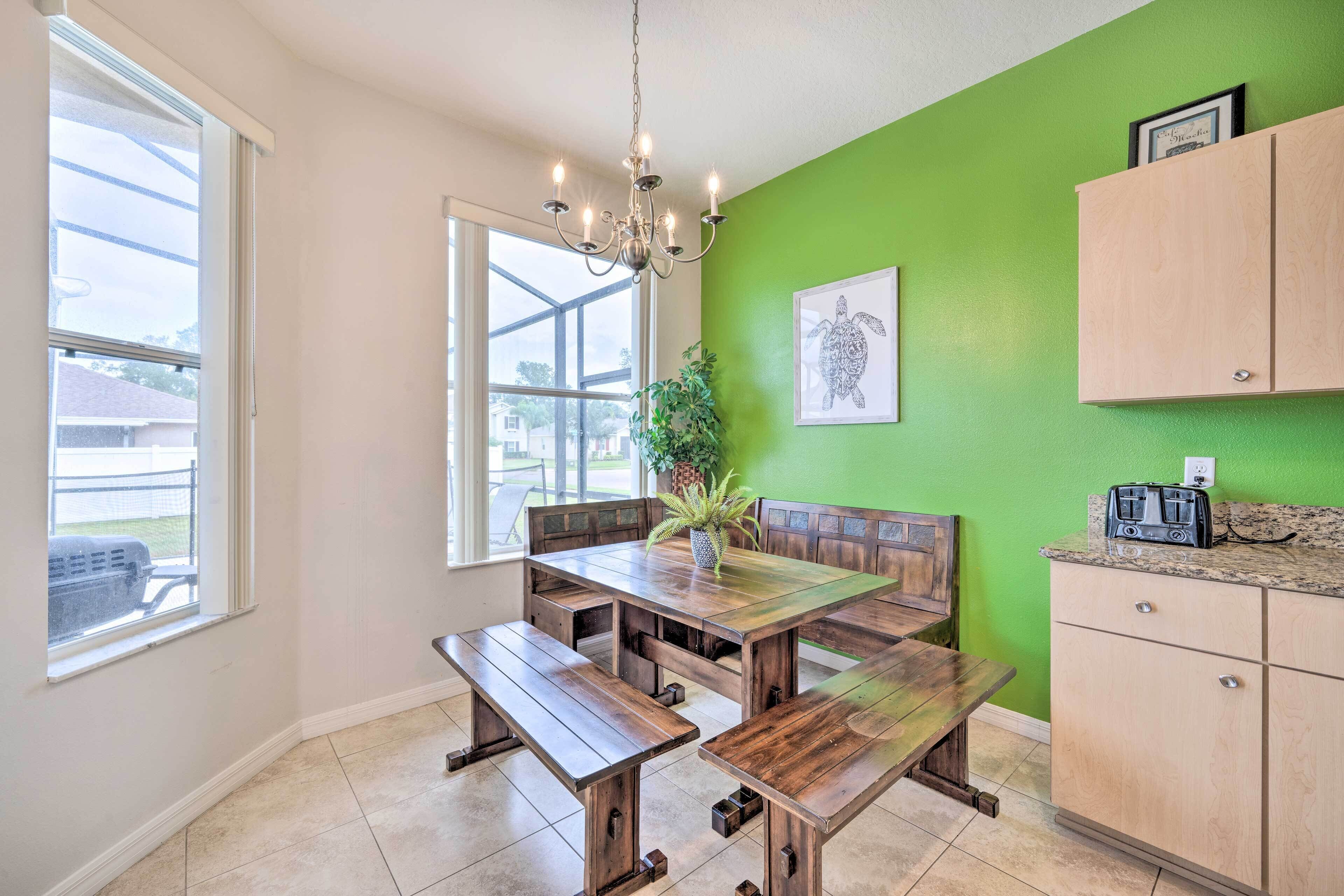 A dining area with a wooden table and benches, a chandelier, a green wall with a framed picture, and a kitchen area with cabinets and a toaster.