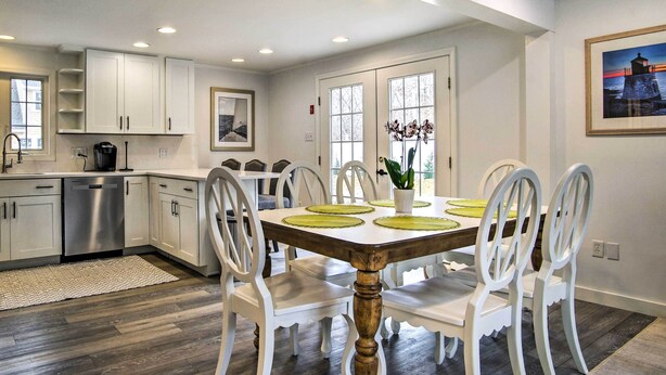 A modern kitchen with white cabinetry, a stainless steel dishwasher, and a dining area with a wooden table and white chairs. - Martha's Vineyard Escape Near South Beach (Edgartown)