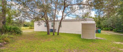 A single-story mobile home with a small attached garage, surrounded by a grassy yard and trees.