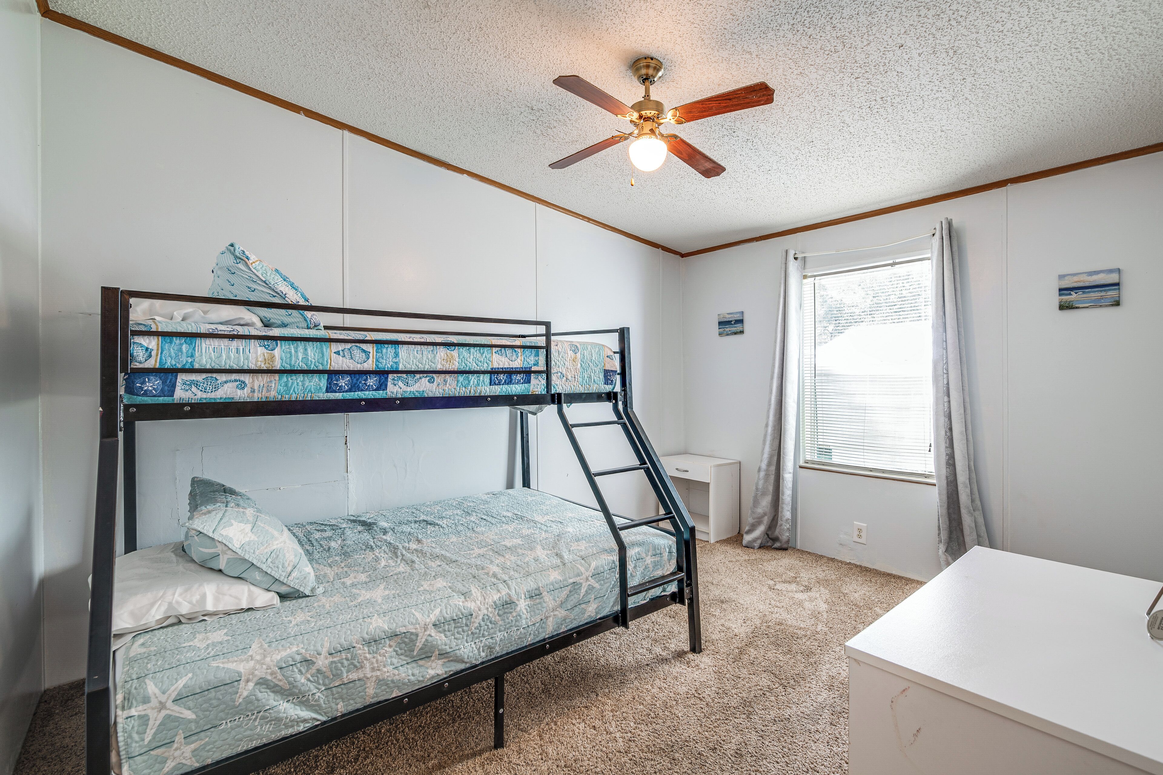 Un lit superposé avec un matelas orné d’un motif d’étoile de mer, un ventilateur de plafond, une fenêtre avec des rideaux et un petit bureau.