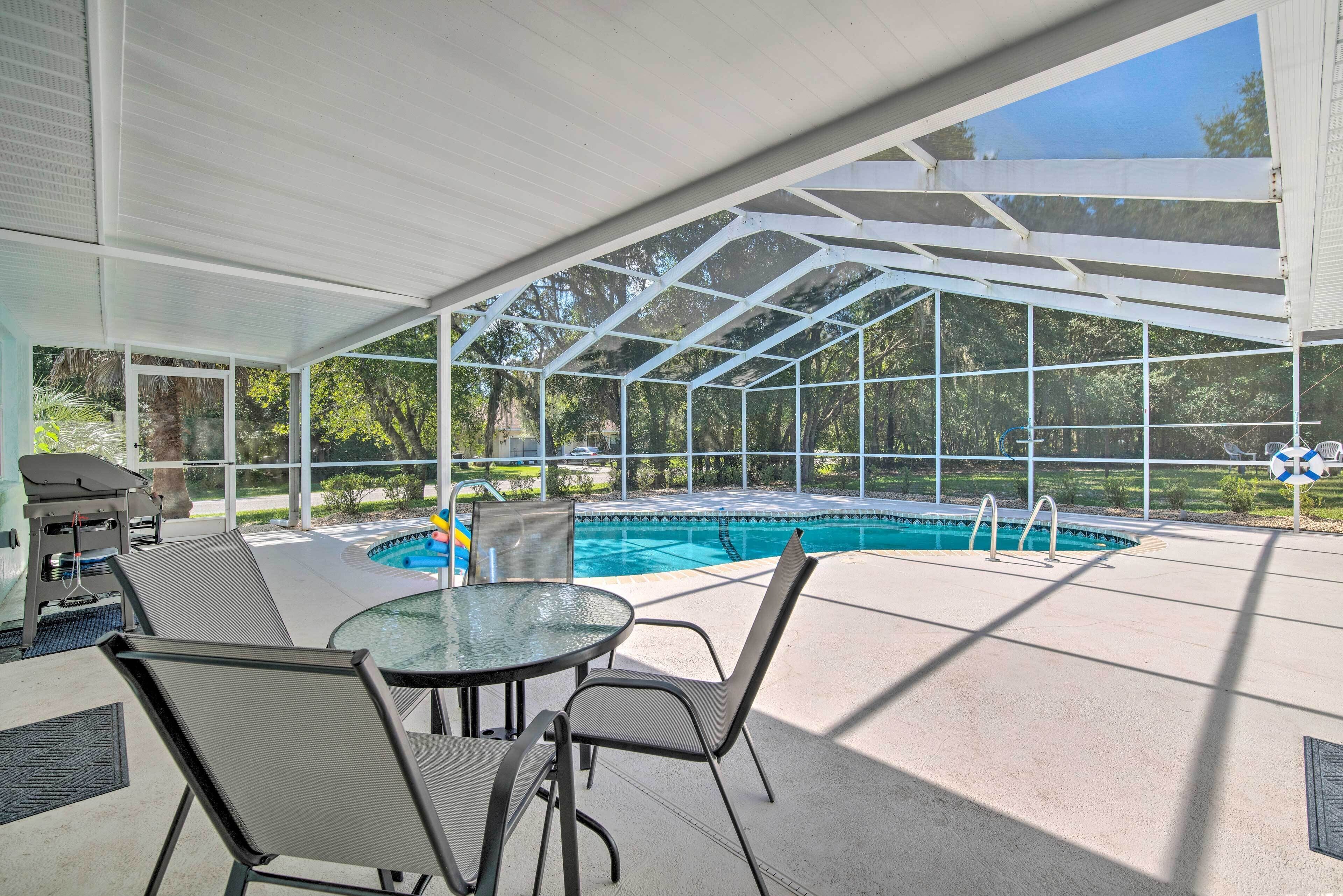 An outdoor pool area with a covered patio, a table with chairs, and a barbecue grill.