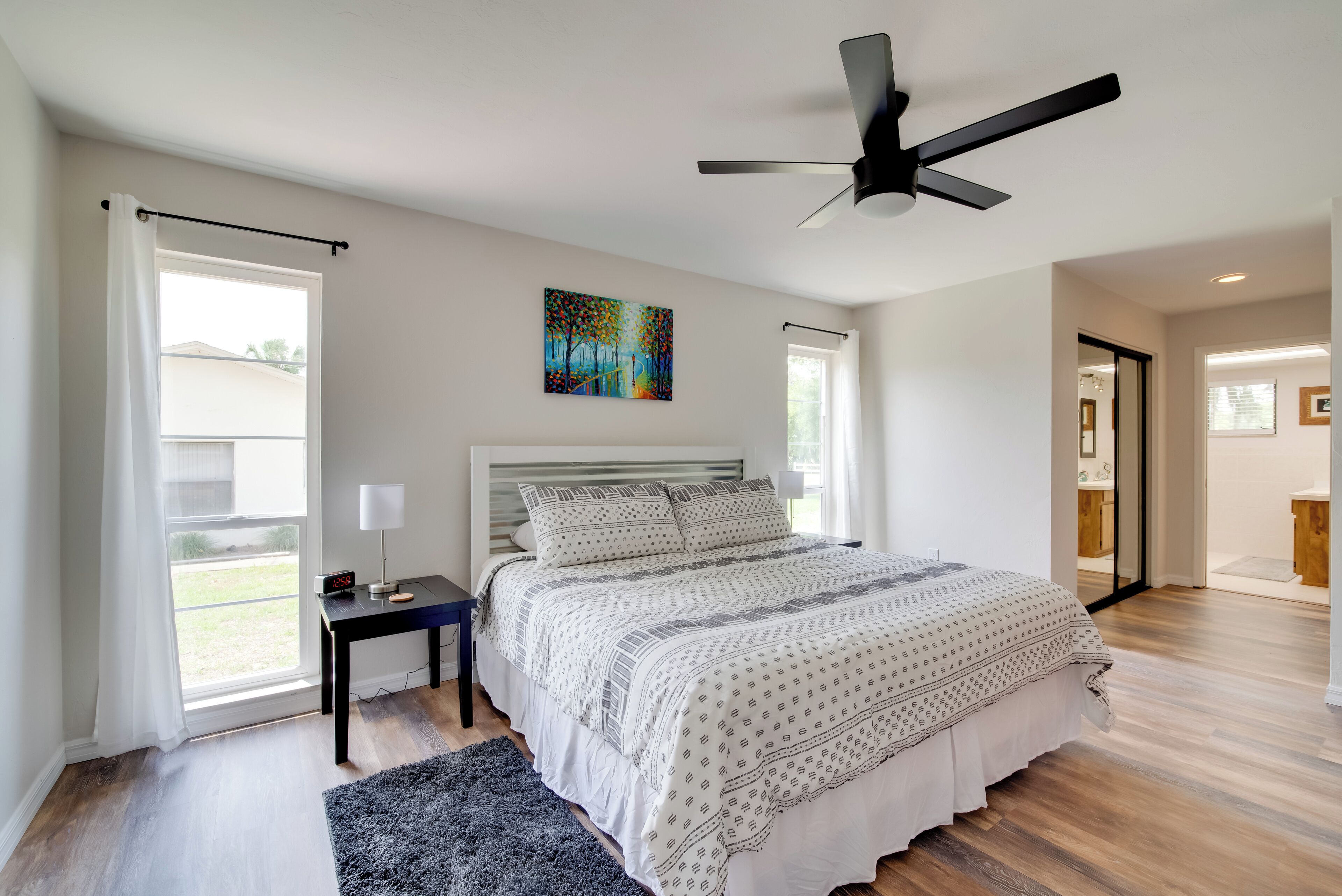 A modern bedroom with a large bed, a ceiling fan, a painting, and a view of the outdoors.