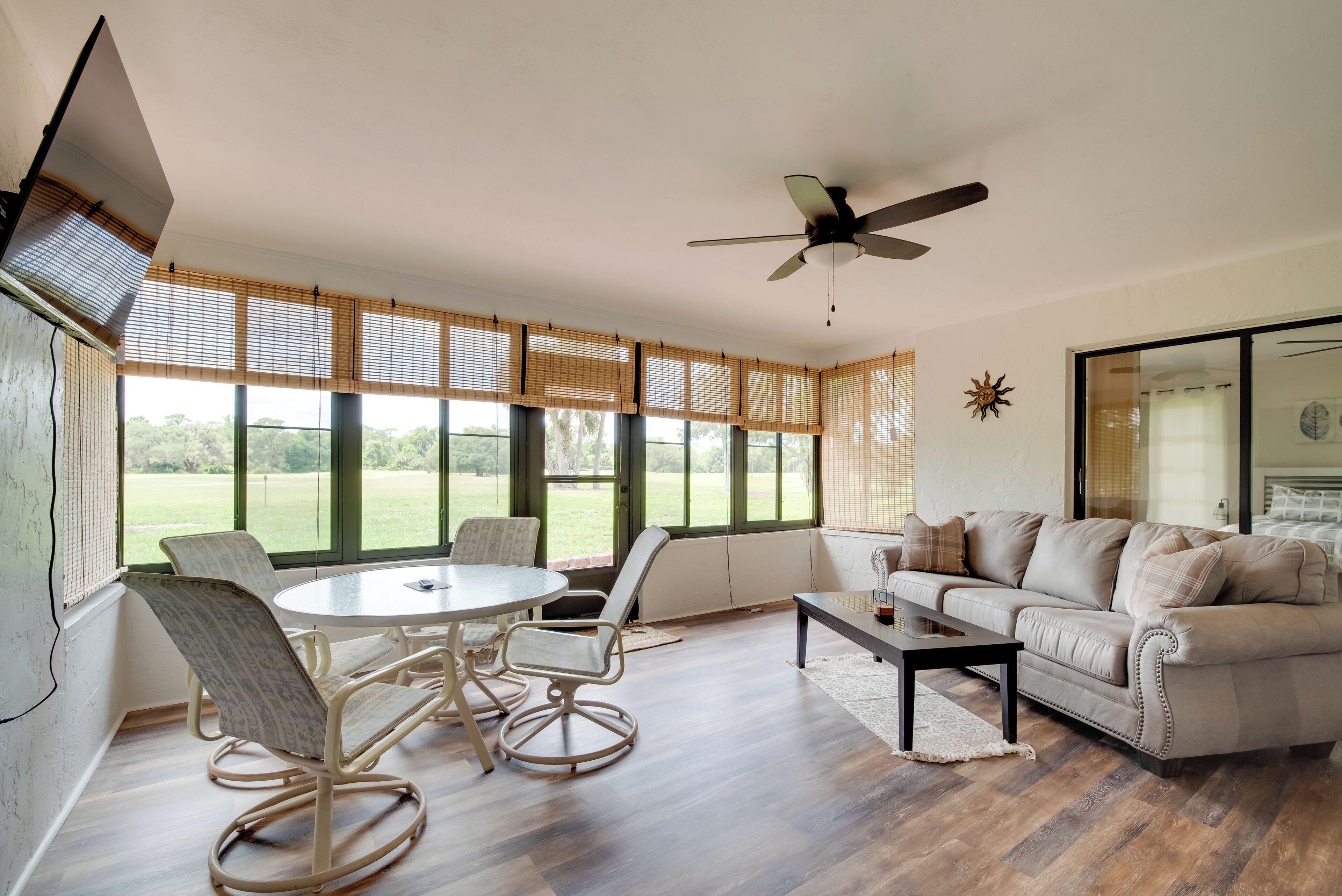 A spacious living room with a round table, two rocking chairs, a beige sofa, and large windows with blinds.