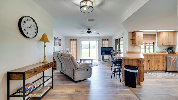 A spacious living area with a sofa, a coffee table, and a dining area with bar stools. There is a kitchen with wooden cabinets and a built-in oven. A ceiling fan and a clock are visible on the wall. - Walk to Lake: Geataway w/ Yard in Inverness (Inverness)