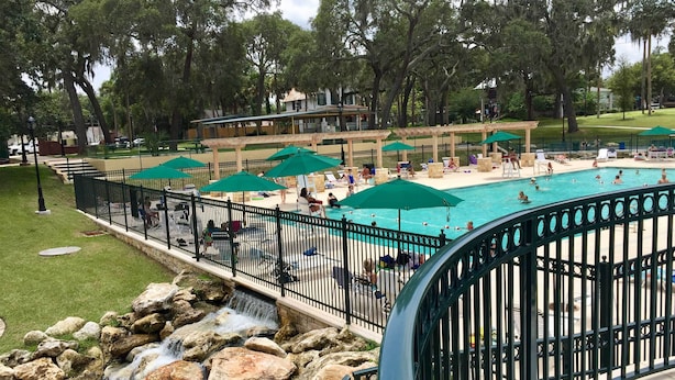 A swimming pool with a waterfall feature, surrounded by a fence and shaded by umbrellas. - Historic Green Cove Springs Home - Walk to Coast! (Green Cove Springs)