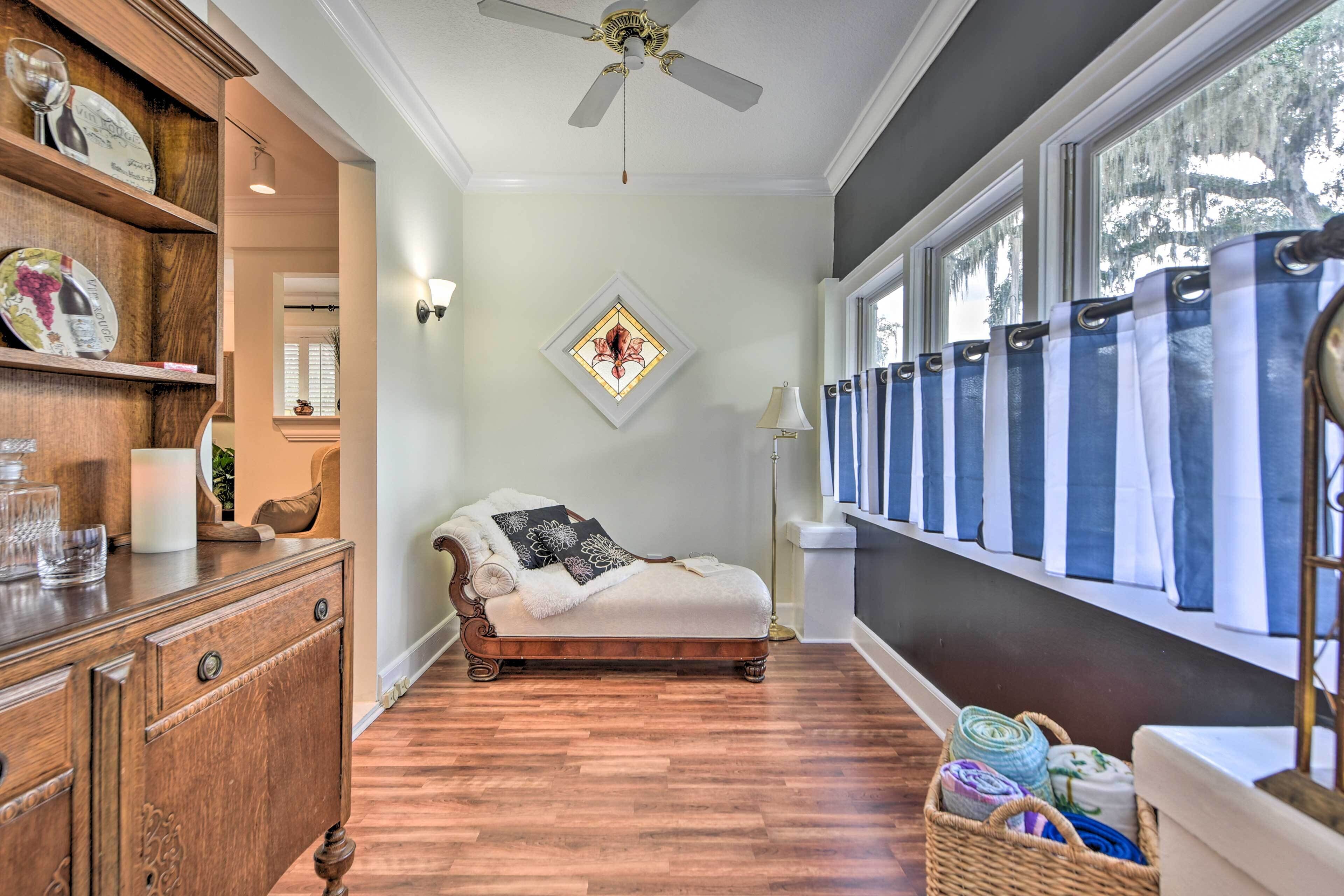 A room with a wooden floor, a bed with a white cover, a ceiling fan, a window with curtains, and a wall-mounted light fixture.