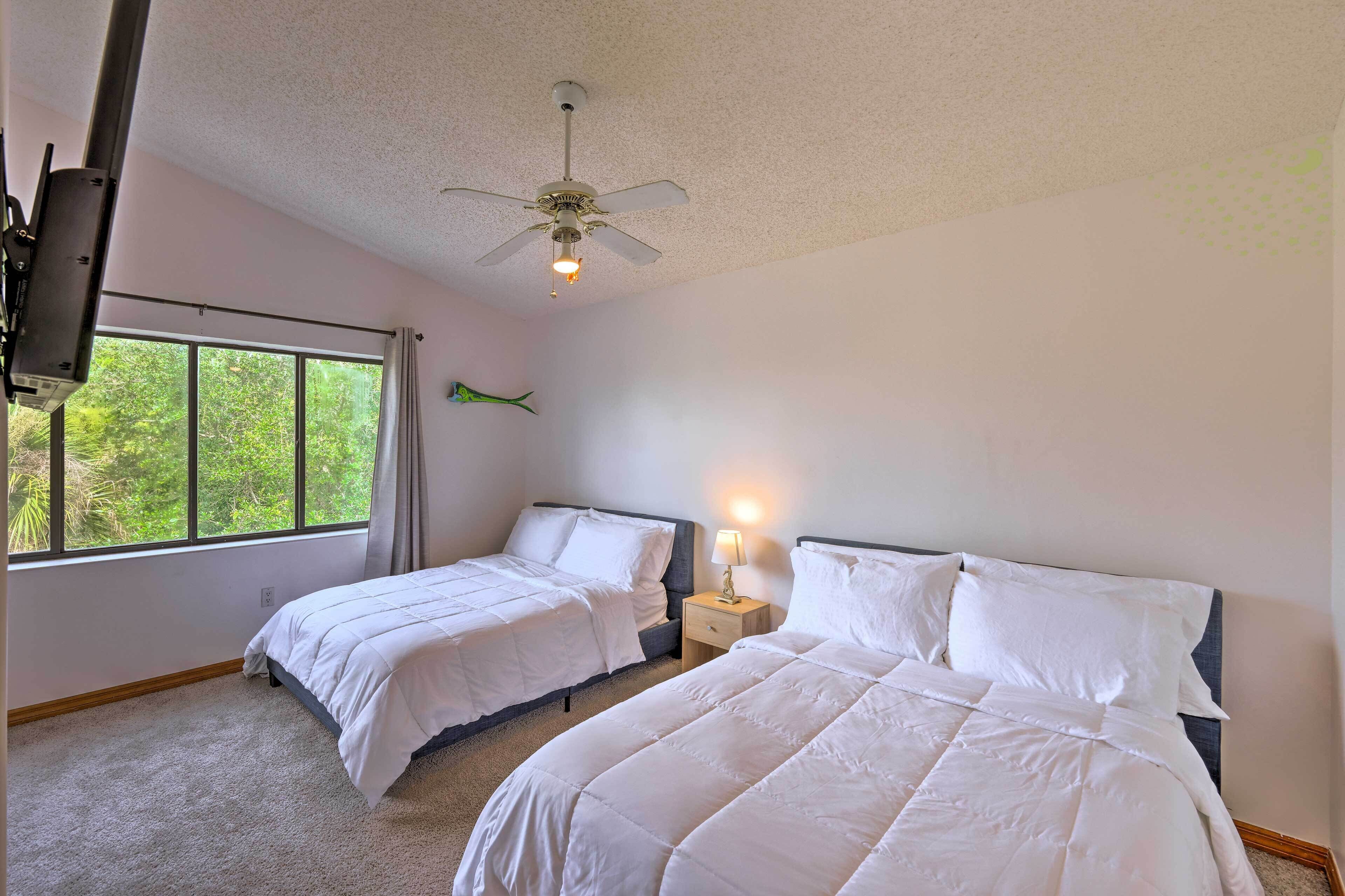 A bedroom with two beds, a ceiling fan, a nightstand with a lamp, and a large window with a view of greenery.