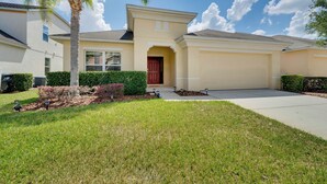 A modern two-story house with a red door, a covered driveway, and well-maintained landscaping including a palm tree and shrubs. - Central Family Home w/ Lanai < 10 Mi to Disney! (Davenport)