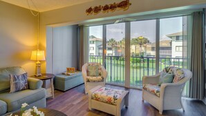 A spacious living room with a sofa, two armchairs, a coffee table, and a lamp. A large sliding glass door leads to a balcony with a view of the neighborhood. - Sunny Ocean Village Beach Condo: Community Pool (Fort Pierce)