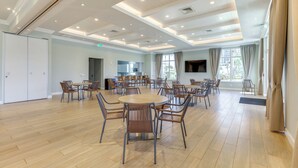 A spacious room with multiple round tables and chairs, a kitchen area, and large windows with curtains. - Sunny Ocean Village Beach Condo: Community Pool (Fort Pierce)