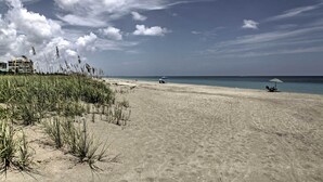 A beach with sand, sparse vegetation, and a distant umbrella. - Sunny Ocean Village Beach Condo: Community Pool (Fort Pierce)