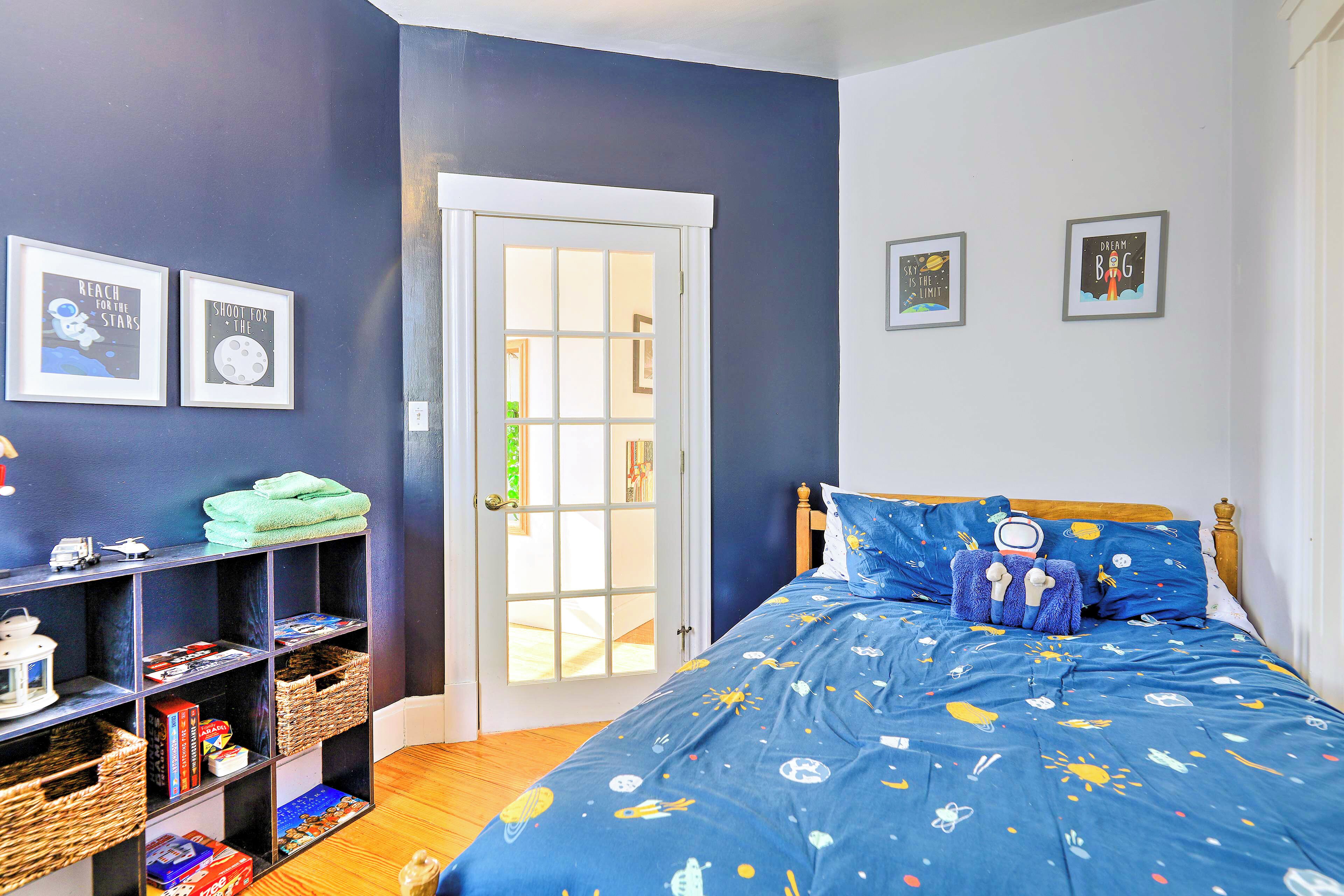 A bedroom with a blue space-themed bedspread, a wooden headboard, and a bookshelf with various items.