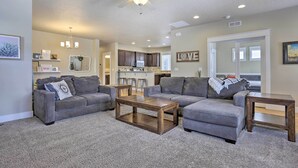 A living room with a gray sectional sofa, a wooden coffee table, and a gray carpet. There is a ceiling fan, recessed lighting, and a kitchen in the background. - Charming Boise Home ~ 8 Mi to Downtown! (Garden City)