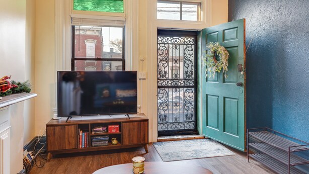 A living room with a television on a wooden stand, a green door with a decorative wreath, and a window with a view of buildings. - Cincinnati Retreat w/ Patio & Grill! (Cincinnati)