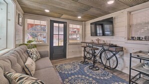 A cozy living room with a sofa, a coffee table, and a patterned rug. There is a kitchen area with a stove, a sink, and a window with blinds. - Peaceful Cookeville Cabin in Unique Setting (Cookeville)