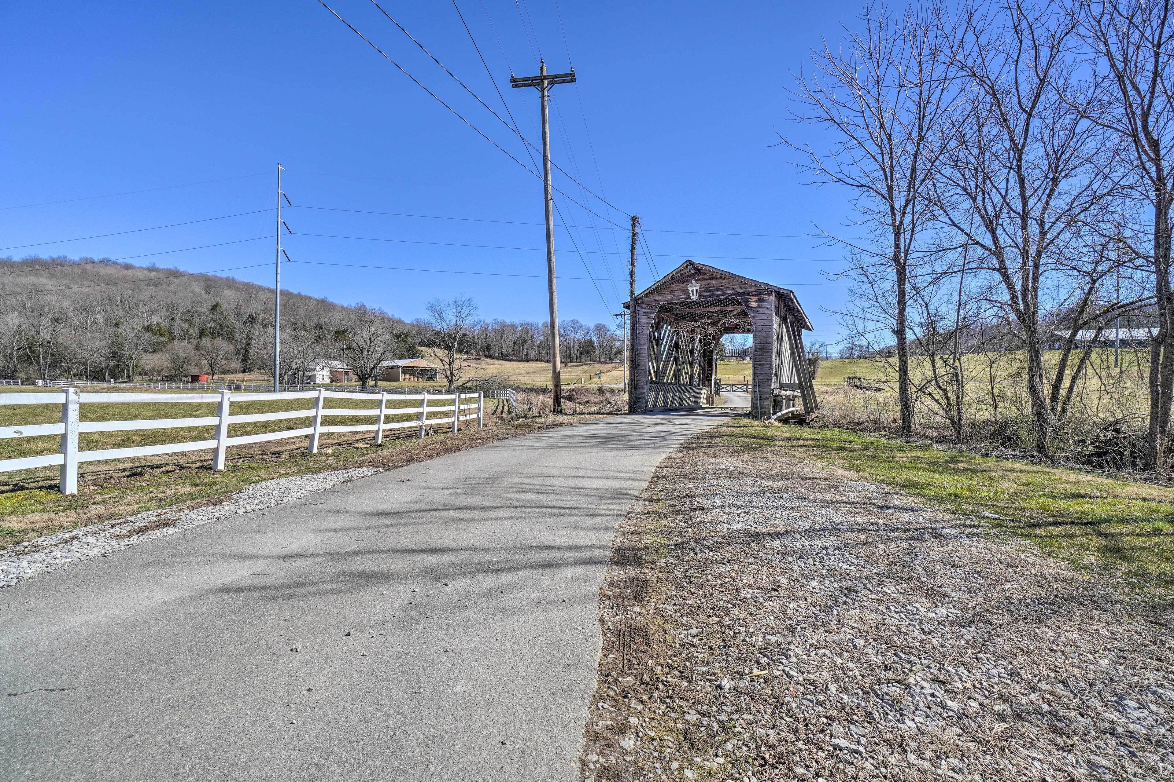 A covered bridge with a gravel path leading to it, surrounded by trees and a white fence.