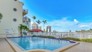 A swimming pool with a white building, palm trees, and a city skyline in the background. - Fort Lauderdale Vacation Rental: Walk to Beach! (Fort Lauderdale)