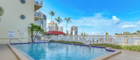 A swimming pool with a white building, palm trees, and a city skyline in the background.