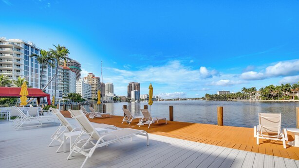 A waterfront deck with white lounge chairs and yellow umbrellas, overlooking a calm body of water and a city skyline. - Fort Lauderdale Vacation Rental: Walk to Beach! (Fort Lauderdale)