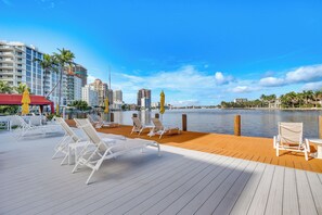 A waterfront deck with white lounge chairs and yellow umbrellas, overlooking a calm body of water and a city skyline. - Fort Lauderdale Vacation Rental: Walk to Beach! (Fort Lauderdale)