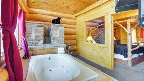 A log cabin room with a stone fireplace, a wooden bed, a large bathtub, and a window with a view of the outside. - Gatlinburg Vacation Rental w/ Hot Tub & Game Room! (Gatlinburg)