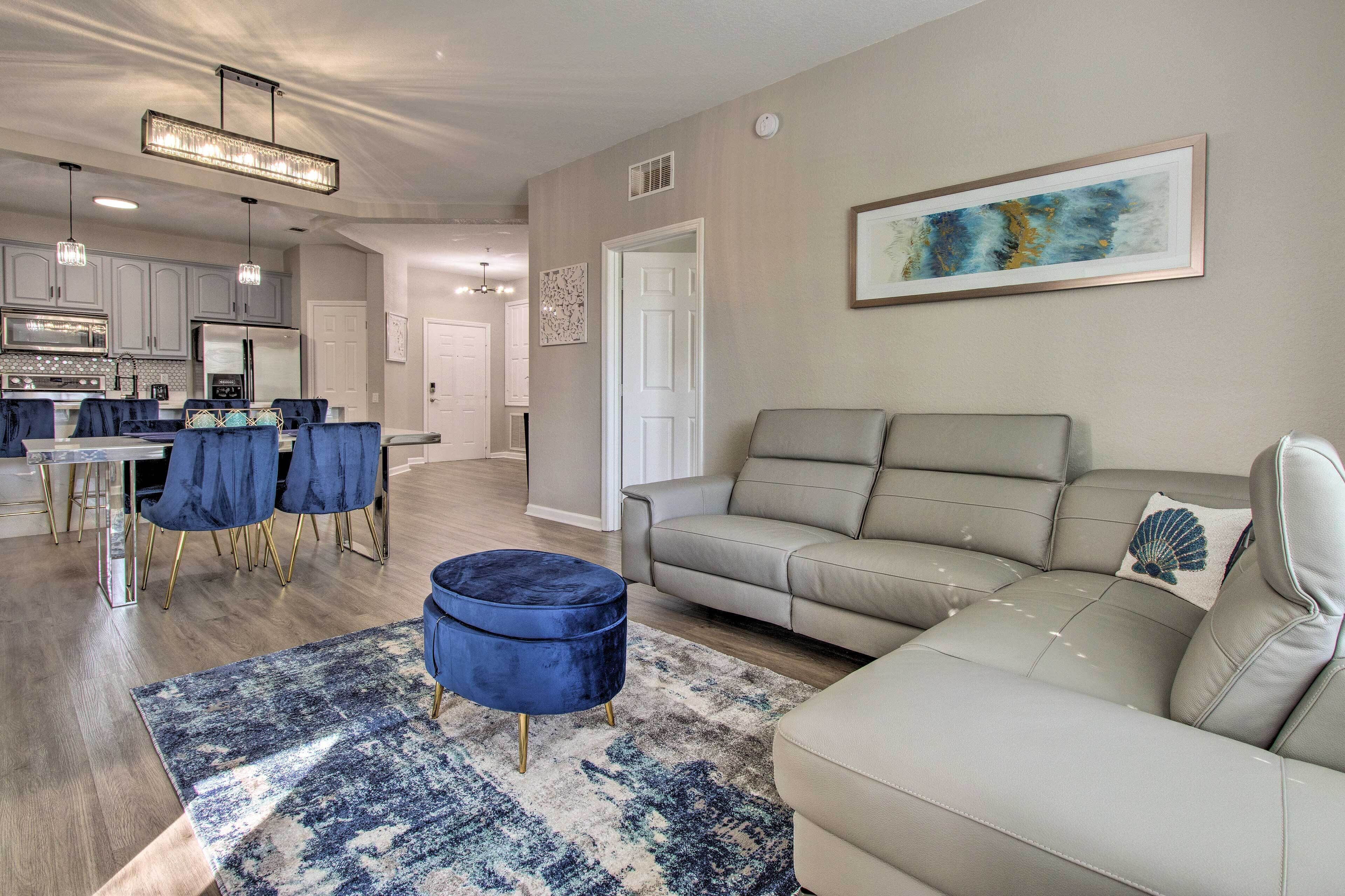 Un salon moderne avec un canapé beige, un pouf bleu et un coin repas comprenant des chaises bleues et un lustre.