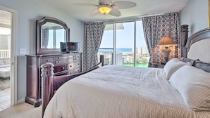 A bedroom with a large bed, a dresser, a mirror, a ceiling fan, and a view of the ocean. - The Terrace Beach View at Pelican Beach Resort (Destin)
