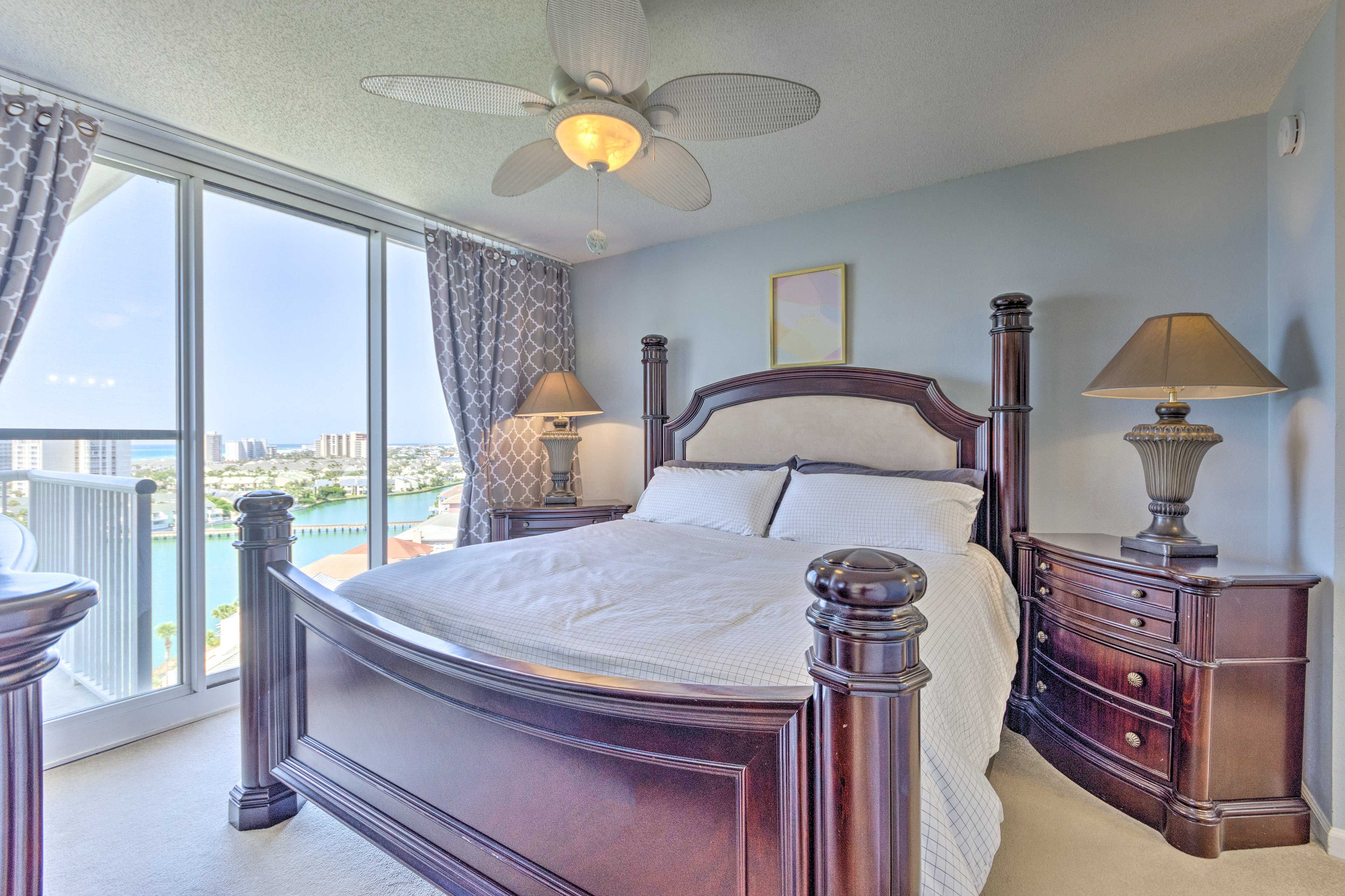 Une chambre à coucher avec un grand lit, deux tables de chevet avec des lampes, un ventilateur de plafond et une vue sur le paysage urbain à travers de grandes fenêtres.