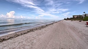 Una playa con cielo despejado y mar tranquilo. Se ven edificios y palmeras al fondo.
