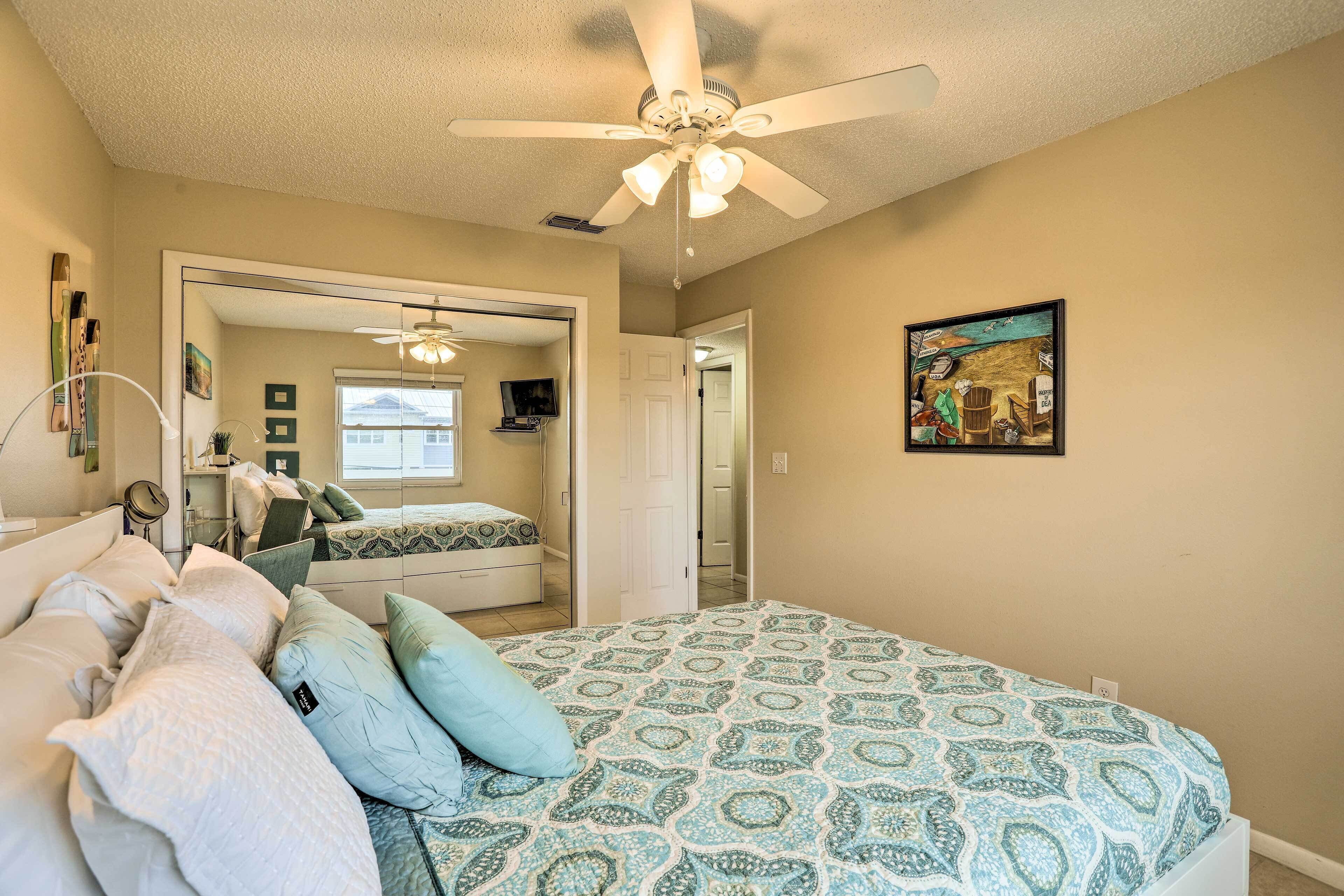 A bedroom with a bed, a ceiling fan, a painting, and a door leading to another room.