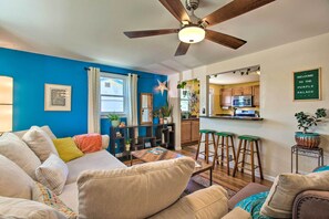A living room with a blue wall, a ceiling fan, a sofa, a coffee table, and a kitchen area with a bar stool. - Patio, Grill & Yard: Cozy Denver Abode! (Denver)