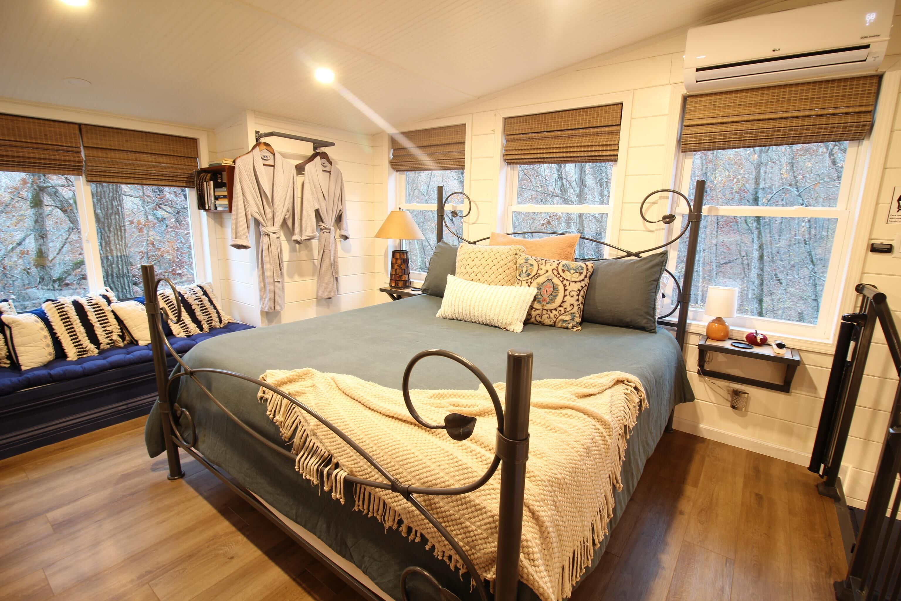 A bedroom with a large bed, a bedside table with a lamp, a hanging robe, and a window with a view of trees.