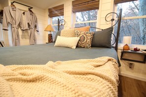 A bedroom with a bed, pillows, a bedside table, a lamp, and a window with a view of trees.
