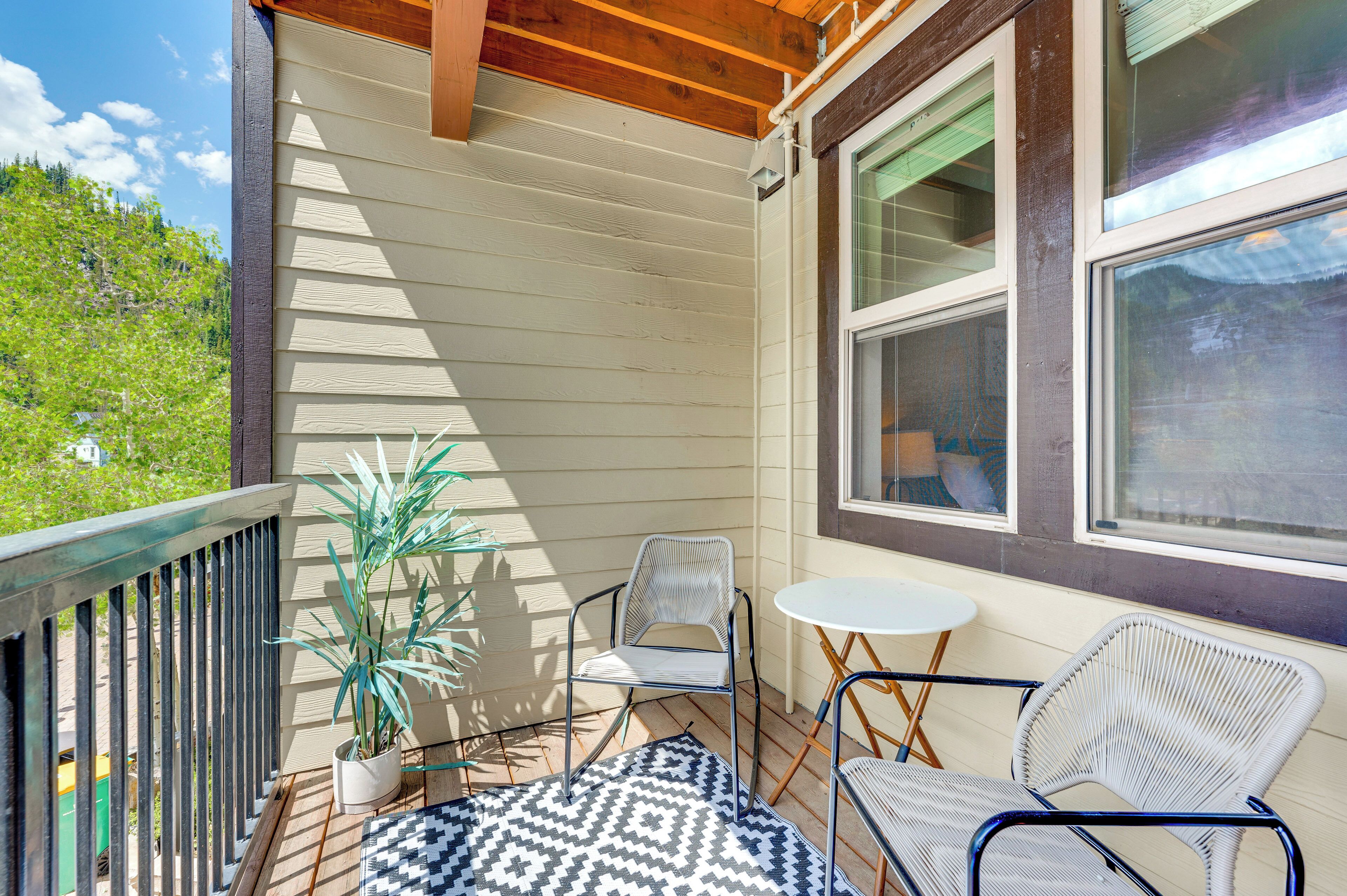 A covered outdoor seating area with a table and two chairs, a potted plant, and a patterned rug.