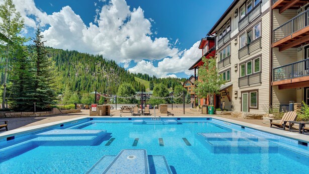 A clear blue pool with multiple lanes, surrounded by a building with balconies and a forested hillside in the background.
