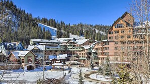 A ski resort with multiple buildings, including a large lodge and smaller cabins, surrounded by snow-covered trees and a clear blue sky.