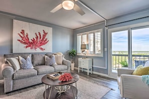 A living room with a gray sofa, a coffee table, a ceiling fan, a large window with blinds, and a balcony view. - PCB Beachfront 'coastal Abode' at Pinnacle Port! (Panama City Beach)